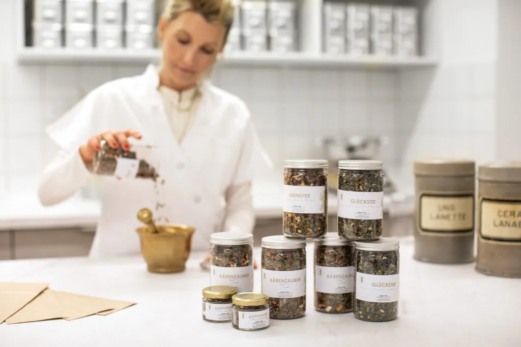 Apothekerin im weißen Mantel füllt Kräuter aus Glasbehältern in einen Messingmörser, daneben Teemischungen der Eigenmarke